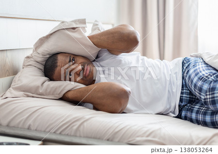 Young african american man lying in bed covering ears with pillow Young african american man lying in bed covering ears with pillow 138053264
