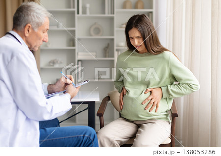 Pregnant woman holding stomach during medical consultation while doctor writes notes 138053286