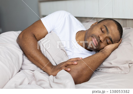 Young black man sleeping peacefully on bed under blanket in cozy bedroom Young black man sleeping peacefully on bed under blanket in cozy bedroom 138053382