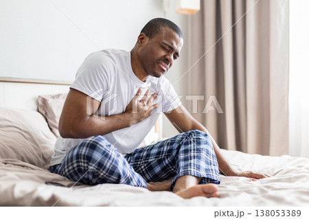 Young black man sitting on bed holding chest in pain, showing discomfort Young black man sitting on bed holding chest in pain, showing discomfort 138053389