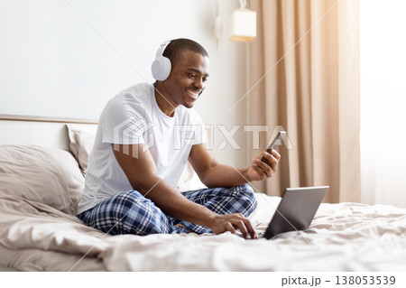 Young black man sitting on bed using smartphone and laptop while smiling Young black man sitting on bed using smartphone and laptop while smiling 138053539
