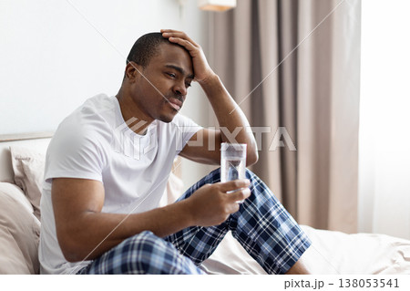 Black man sitting on bed holding glass of water and touching head Black man sitting on bed holding glass of water and touching head 138053541