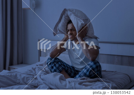 Young black man sitting in bed at night covering ears with pillow 138053542