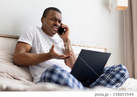 Worry black man sitting on bed talking on smartphone while using laptop 138053544