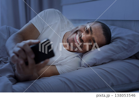 Young black man lying in bed at night smiling while using smartphone Young black man lying in bed at night smiling while using smartphone 138053546