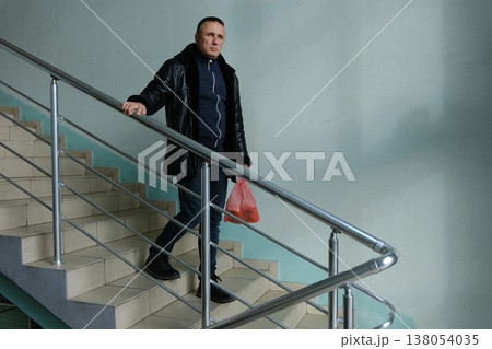 Middle-aged man examining shopping red plastic bag standing on steps leather jacket casual outdoor lifestyle groceries urban scene Middle-aged man examining shopping red plastic bag standing on steps leather jacket casual outdoor lifestyle groceries urban scene 138054035