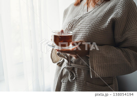 Woman in bathrobe drinking hot tea by window, finding peace and relaxation at home during a tranquil morning routine Woman in bathrobe drinking hot tea by window, finding peace and relaxation at home during a tranquil morning routine 138054194