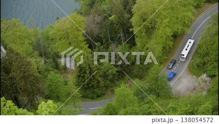 Cars travel on a winding road near a lake surrounded by green trees during the day in a rural area 138054572