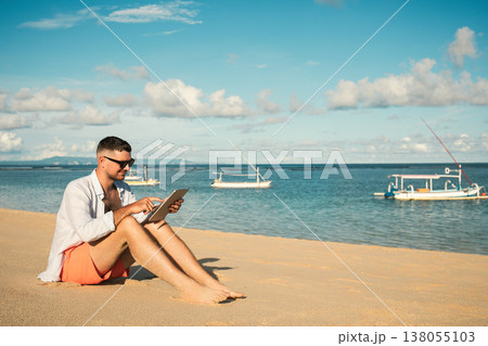 Man sits on the sand using a tablet near the water 138055103