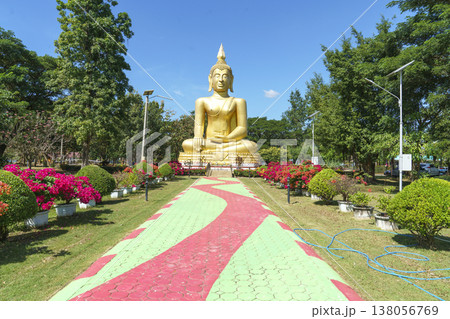 Large golden Buddha statue seated in a beautiful public park with colorful flowers and green trees under a clear blue sky. A wavy patterned path leads toward the majestic religious landmark. 138056769