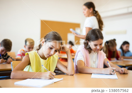 Interested preteen girl studying in classroom with classmates Interested preteen girl studying in classroom with classmates 138057562