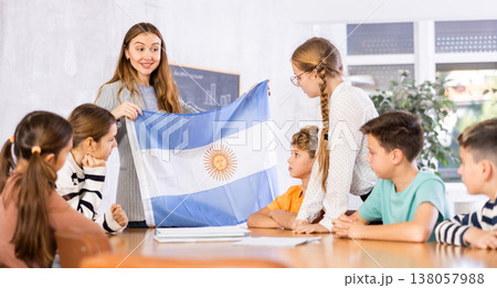 Female teacher, conducting extracurricular lesson in the school class tells schoolchildren the history of Argentina and shows national flag of country 138057988