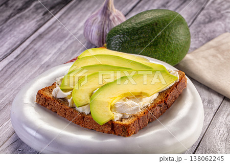 Avocado toast with soft cheese on dark rye bread. A healthy breakfast sandwich on a white plate over a rustic wooden table. 138062245