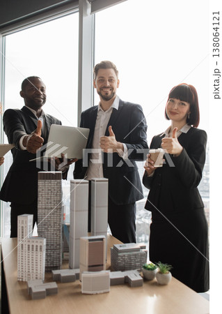 A diverse group of professionals in business attire gives a thumbs-up gesture while standing near architectural models and a laptop 138064121