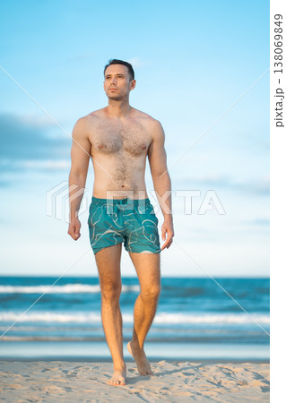 handsome white man with athletic muscular body standing at beach at sunny sunset, attractive male athlete walking at sea or ocean shore 138069849