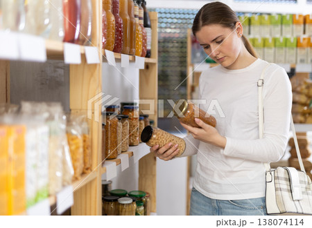 Girl customer in supermarket examines jar of kidney bean and lentil, chooses food-stuff in grocery Girl customer in supermarket examines jar of kidney bean and lentil, chooses food-stuff in grocery 138074114