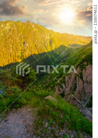 sunset over valley in summer mountains. Gorgeous mountainous landscape. Rocky cliffs and forested slopes under cloudy sky. Scenery of mountain ridge in evening light. sunny weather with rainbow 138074611