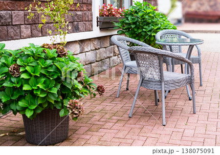 Outdoor cafe patio with wicker furniture set including table and chair on stone paved ground. Cozy restaurant terrace with green plant in pot near brick wall for exterior design concept. Outdoor cafe patio with wicker furniture set including table and chair on stone paved ground. Cozy restaurant terrace with green plant in pot near brick wall for exterior design concept. 138075091