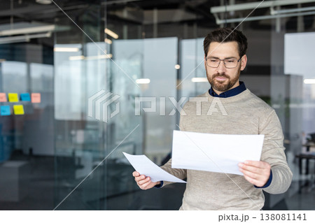 Young businessman standing in a contemporary glass office, reading and analyzing financial documents with a focused expression, representing data review and corporate management Young businessman standing in a contemporary glass office, reading and analyzing financial documents with a focused expression, representing data review and corporate management 138081141