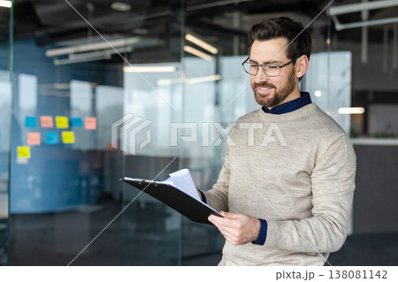 Young businessman smiling while carefully examining documents on a clipboard, standing in a modern office environment with glass walls and sticky notes Young businessman smiling while carefully examining documents on a clipboard, standing in a modern office environment with glass walls and sticky notes 138081142
