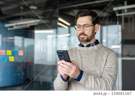 Man in an open-plan office setting, wearing eyeglasses and a sweater, intently using a smartphone in his hands for remote work, social media, or other digital communication 138081167