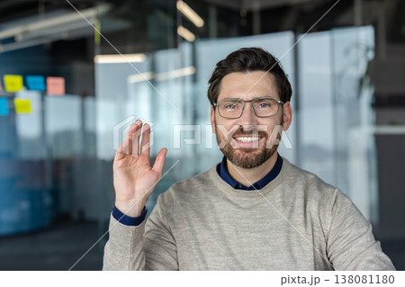 Caucasian man with glasses and a beard smiling and waving his hand, participating in a video conference or online meeting from a modern office environment Caucasian man with glasses and a beard smiling and waving his hand, participating in a video conference or online meeting from a modern office environment 138081180