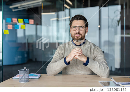 Confident businessman wearing eyeglasses and a sweater, sitting at a desk with his hands clasped, working in a contemporary office setting with sticky notes on a glass wall Confident businessman wearing eyeglasses and a sweater, sitting at a desk with his hands clasped, working in a contemporary office setting with sticky notes on a glass wall 138081183