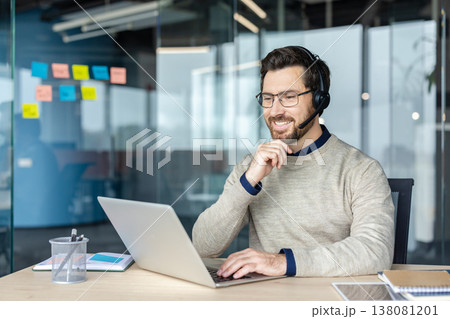 Smiling man wearing a headset and spectacles working on his laptop, providing online customer support or attending a virtual meeting from a modern glass-walled office workspace Smiling man wearing a headset and spectacles working on his laptop, providing online customer support or attending a virtual meeting from a modern glass-walled office workspace 138081201