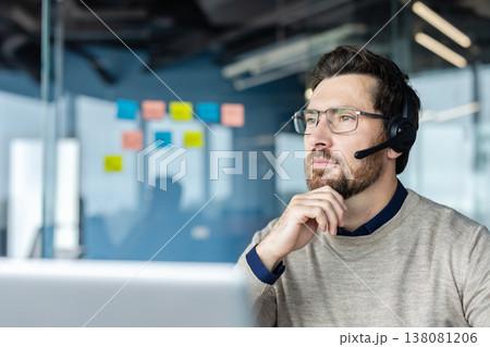 Bearded call center agent with glasses and headset in a modern office, looking away thoughtfully while providing customer support and technical assistance with concentration and professionalism 138081206
