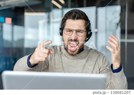 Man wearing a headset and glasses, expressing extreme anger and frustration, pointing his finger with an open mouth while working at a laptop in a modern office setup Man wearing a headset and glasses, expressing extreme anger and frustration, pointing his finger with an open mouth while working at a laptop in a modern office setup 138081207