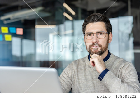 Man with glasses and beard pausing at his laptop in a contemporary office, hand on chin, focused and thoughtful while planning strategy and considering solutions for business success Man with glasses and beard pausing at his laptop in a contemporary office, hand on chin, focused and thoughtful while planning strategy and considering solutions for business success 138081222
