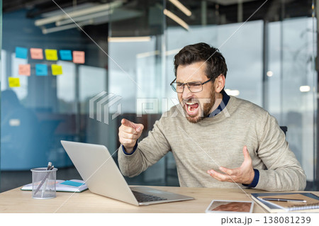Stressed business man shouting and pointing at his laptop screen in a modern office, furious and overwhelmed by technology failure, deadlines and workplace pressure Stressed business man shouting and pointing at his laptop screen in a modern office, furious and overwhelmed by technology failure, deadlines and workplace pressure 138081239