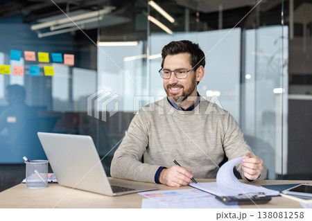 Smiling man in eyeglasses sitting at a desk, looking at his laptop screen while holding a pen and flipping through papers, focusing on his work in a modern office 138081258