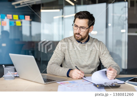 Professional man wearing glasses working on a laptop at his desk in a modern office, concentrating while reviewing financial documents, project schedules, and data analytics paperwork 138081260