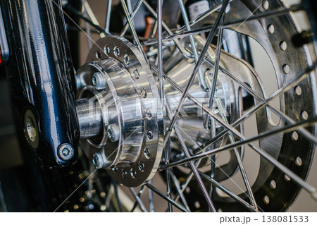 A detailed horizontal macro shot focusing on the polished aluminum front wheel hub, stainless steel wire spokes, and a perforated brake disc of a motorcycle. A detailed horizontal macro shot focusing on the polished aluminum front wheel hub, stainless steel wire spokes, and a perforated brake disc of a motorcycle. 138081533