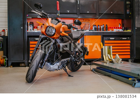 A wide horizontal shot of a sleek orange electric motorcycle parked in a clean, organized garage with black and orange tool cabinets and a hydraulic lift. 138081534