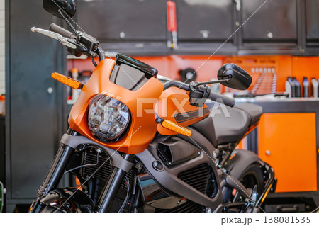 A medium horizontal shot focusing on the front fairing, LED headlight, and handlebars of a matte orange electric motorcycle. Background shows a professional garage setting with orange and black tool A medium horizontal shot focusing on the front fairing, LED headlight, and handlebars of a matte orange electric motorcycle. Background shows a professional garage setting with orange and black tool 138081535