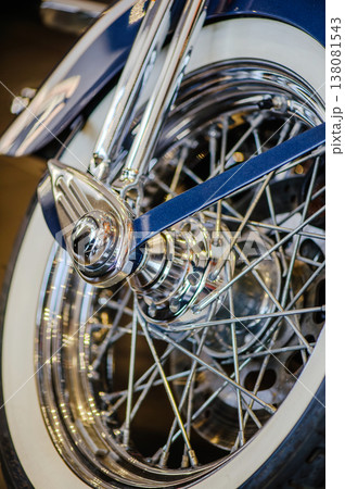 A detailed vertical macro shot of a vintage-style motorcycle's front wheel, showcasing polished chrome wire spokes, a shiny hub, and a classic white-sidewall tire. 138081543