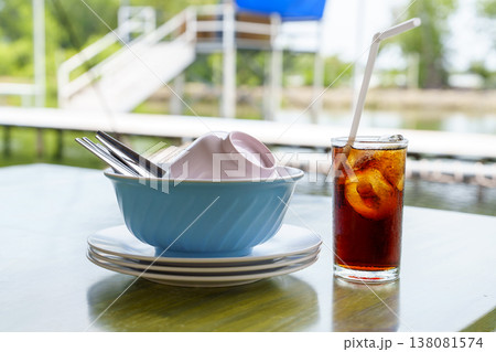Stack of clean ceramic dishes with a blue bowl and plates beside a refreshing glass of iced cola with a straw on an outdoor table. Waterfront restaurant setting with a bright blurred background. Stack of clean ceramic dishes with a blue bowl and plates beside a refreshing glass of iced cola with a straw on an outdoor table. Waterfront restaurant setting with a bright blurred background. 138081574