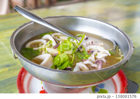Thai spicy squid soup in a stainless steel hot pot served on a colorful tray. The dish features fresh squid rings, aromatic herbs, and lime in a clear broth. Traditional Asian seafood meal. Thai spicy squid soup in a stainless steel hot pot served on a colorful tray. The dish features fresh squid rings, aromatic herbs, and lime in a clear broth. Traditional Asian seafood meal. 138081576