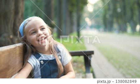 Little happy child girl sitting on a bench smiling happily in summer park. Little happy child girl sitting on a bench smiling happily in summer park. 138082849