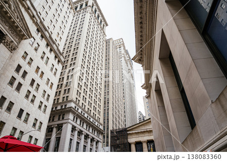 Tall historic skyscrapers rise above Wall Street in Manhattan financial district. The narrow urban canyon highlights classic architecture and modern office towers in downtown New York City. 138083360