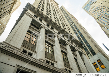 Tall financial skyscraper with classical columns rises above Wall Street in New York City. The architectural facade represents corporate business, banking, and investment institutions in Manhattan. 138083374