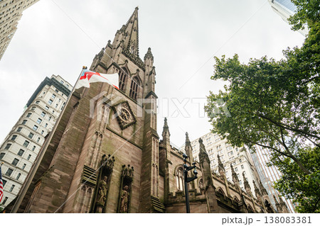 A tall Gothic church spire rises between modern and historic skyscrapers in New York City. The ornate stone architecture contrasts with surrounding office towers in downtown Manhattan. 138083381