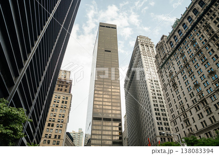 A dramatic low angle view shows tall office towers and skyscrapers in Manhattan New York. Glass and stone buildings reflect light in the busy downtown financial district. 138083394