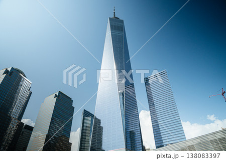 One World Trade Center dominates the Lower Manhattan skyline with reflective glass architecture. The tall tower stands among modern office buildings under a clear blue sky in New York City. 138083397