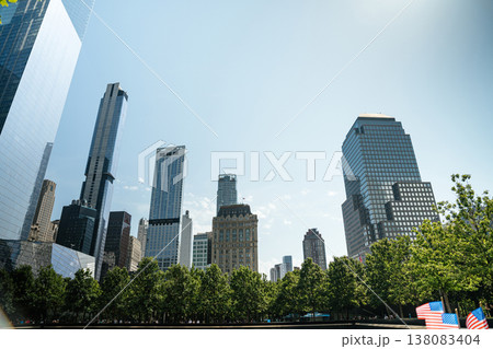 The Lower Manhattan skyline rises behind green trees with an American flag in the foreground. Modern glass skyscrapers stand under a bright blue sky in New York City. The Lower Manhattan skyline rises behind green trees with an American flag in the foreground. Modern glass skyscrapers stand under a bright blue sky in New York City. 138083404