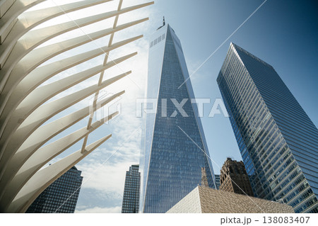 One World Trade Center towers over Lower Manhattan beside the white architectural ribs of the Oculus. The modern glass skyscraper reflects the blue sky in New York City's financial district. One World Trade Center towers over Lower Manhattan beside the white architectural ribs of the Oculus. The modern glass skyscraper reflects the blue sky in New York City's financial district. 138083407