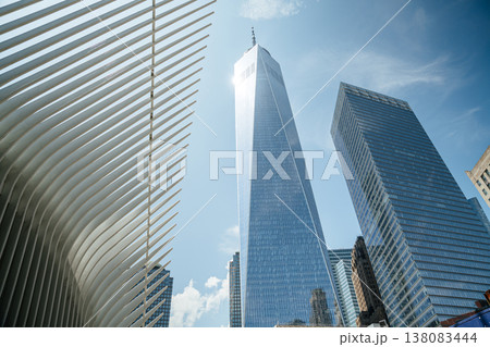 One World Trade Center rises high above downtown New York City next to the white Oculus structure. The glass skyscraper reflects sunlight against a clear blue summer sky. 138083444