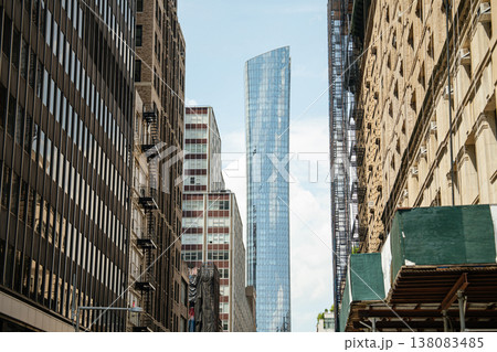 A tall modern glass skyscraper rises between older brick buildings in Manhattan, New York City. The narrow perspective highlights urban density and architectural contrast in downtown NYC. 138083485